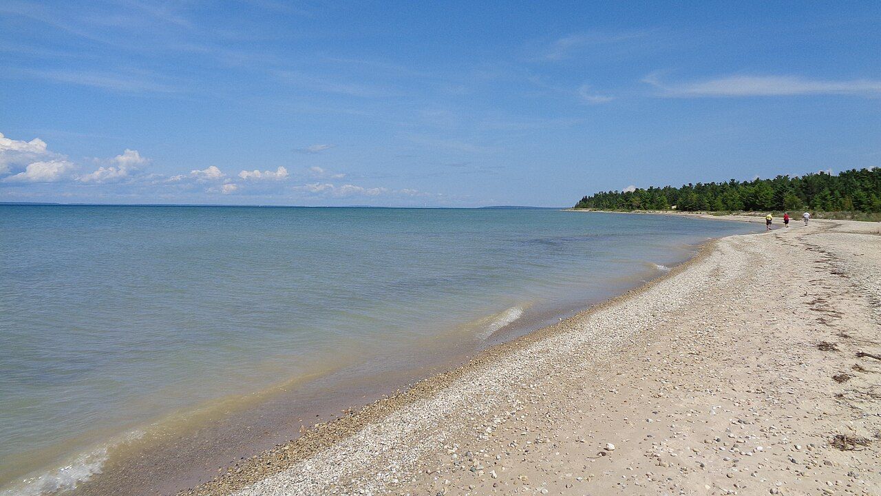 A long pebble beach and calm Lake Michigan water at Wilderness State Park, with woods at the far point.